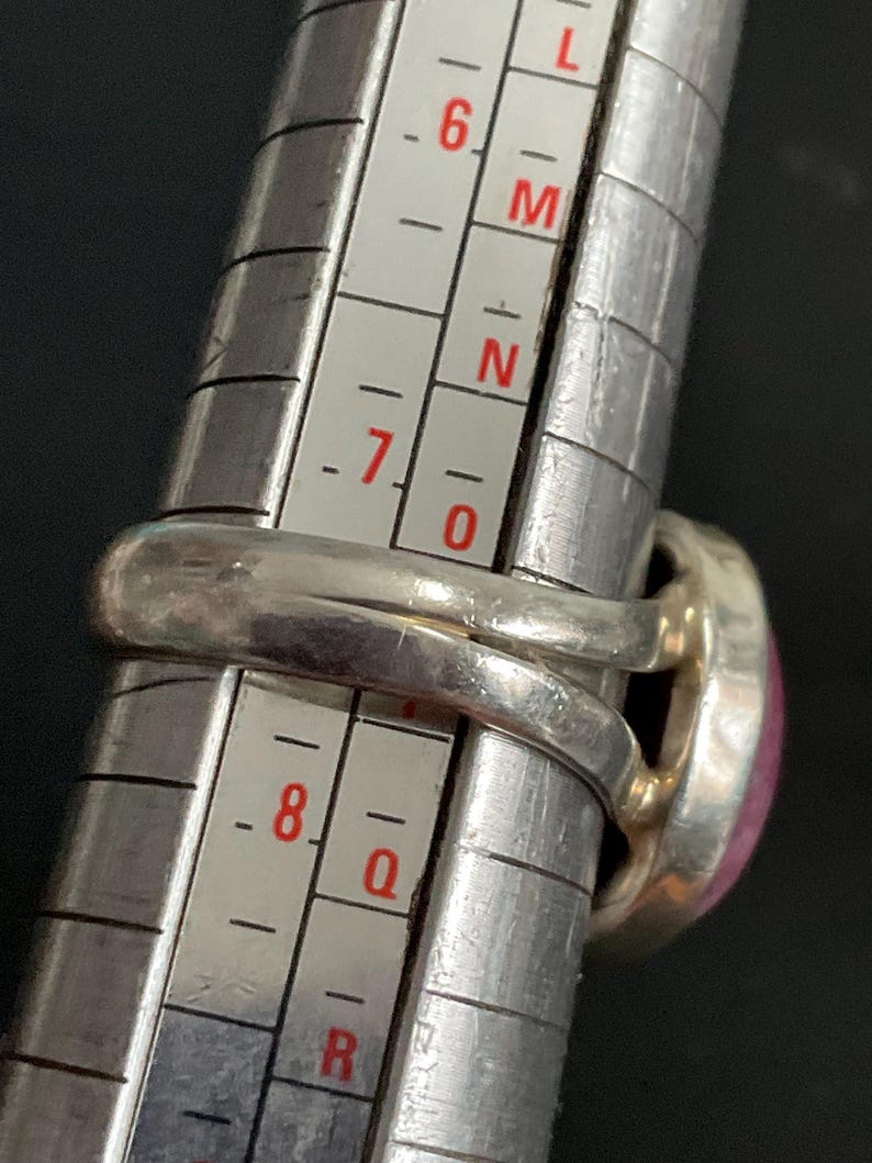 Pu&ograve; includere: Un anello in argento con una pietra rosa posizionato su un mandrino per anelli in metallo. La fascia dell'anello si allinea con la scala di misurazione, che presenta segni numerici e alfabetici per una misurazione precisa della misura.