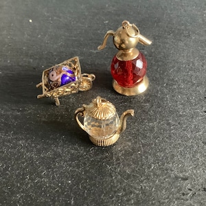 May include: Three gold charm pendants with glass accents. The first charm is a red glass ball with a gold bird-shaped top. The second charm is a clear glass teapot with gold accents. The third charm is a gold wheelbarrow with a blue and white glass ball inside.