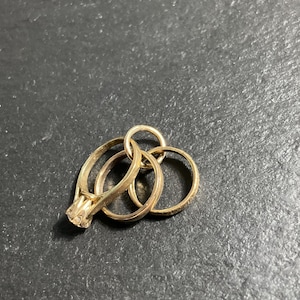 K&ouml;nnte beinhalten: Ein Set aus drei ineinandergreifenden goldfarbenen Ringen, wobei ein Ring an der Oberseite mit einem einzelnen, in Krappen gefassten klaren Stein versehen ist. Die Ringe sind durch einen kleinen runden Verbindungsring zu einem B&uuml;ndel verbunden.