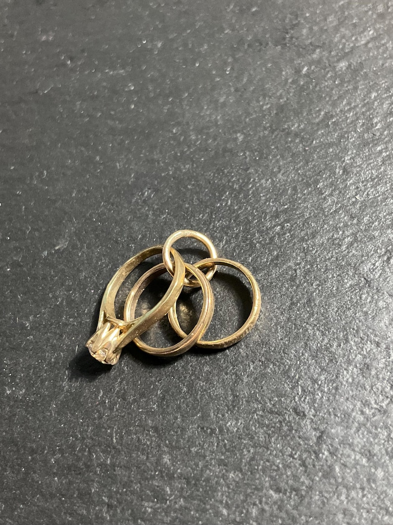 K&ouml;nnte beinhalten: Ein Set aus drei ineinandergreifenden goldfarbenen Ringen, wobei ein Ring an der Oberseite mit einem einzelnen, in Krappen gefassten klaren Stein versehen ist. Die Ringe sind durch einen kleinen runden Verbindungsring zu einem B&uuml;ndel verbunden.