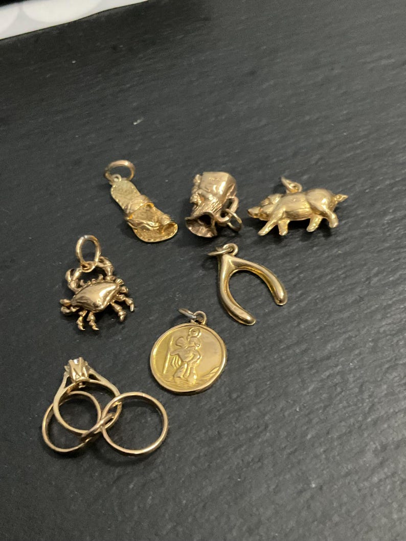 K&ouml;nnte beinhalten: Eine Sammlung goldfarbener Schmuckst&uuml;cke, darunter ein Flip-Flop-Anh&auml;nger, ein kleiner Eimer, eine Schweinefigur, ein Krabbenanh&auml;nger, ein W&uuml;nscheknochen, ein runder Anh&auml;nger mit einer Figur und zwei ineinandergreifende Ringe mit einem Stein.