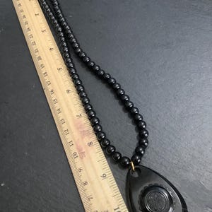 May include: A black beaded necklace with a teardrop pendant. The pendant features a floral design and measures approximately 3 inches in length. The necklace is displayed on a dark surface.