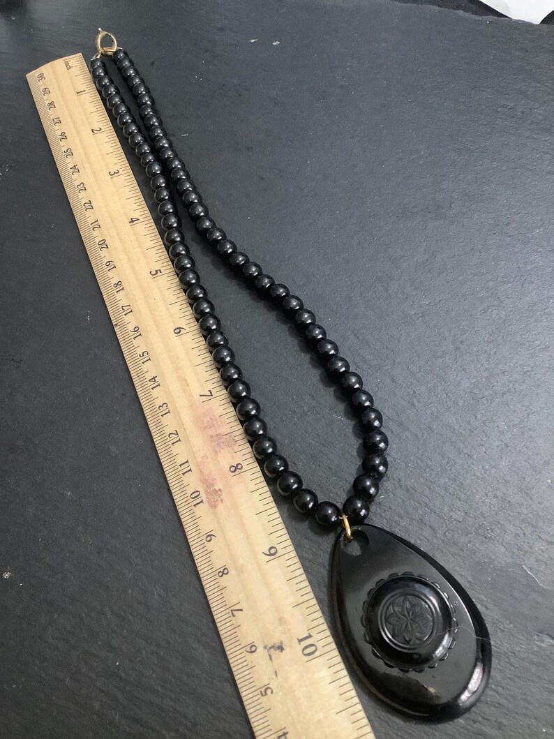 May include: A black beaded necklace with a teardrop pendant. The pendant features a floral design and measures approximately 3 inches in length. The necklace is displayed on a dark surface.