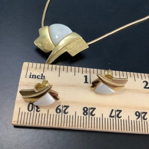 Op de afbeelding: Gouden ketting en oorbellen set met witte opaalstenen. De ketting heeft een ketting en een hanger met een geometrisch ontwerp. De oorbellen zijn klein en hebben een vergelijkbaar ontwerp als de hanger.