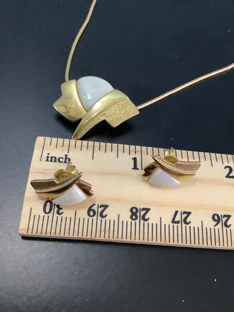 Op de afbeelding: Gouden ketting en oorbellen set met witte opaalstenen. De ketting heeft een ketting en een hanger met een geometrisch ontwerp. De oorbellen zijn klein en hebben een vergelijkbaar ontwerp als de hanger.