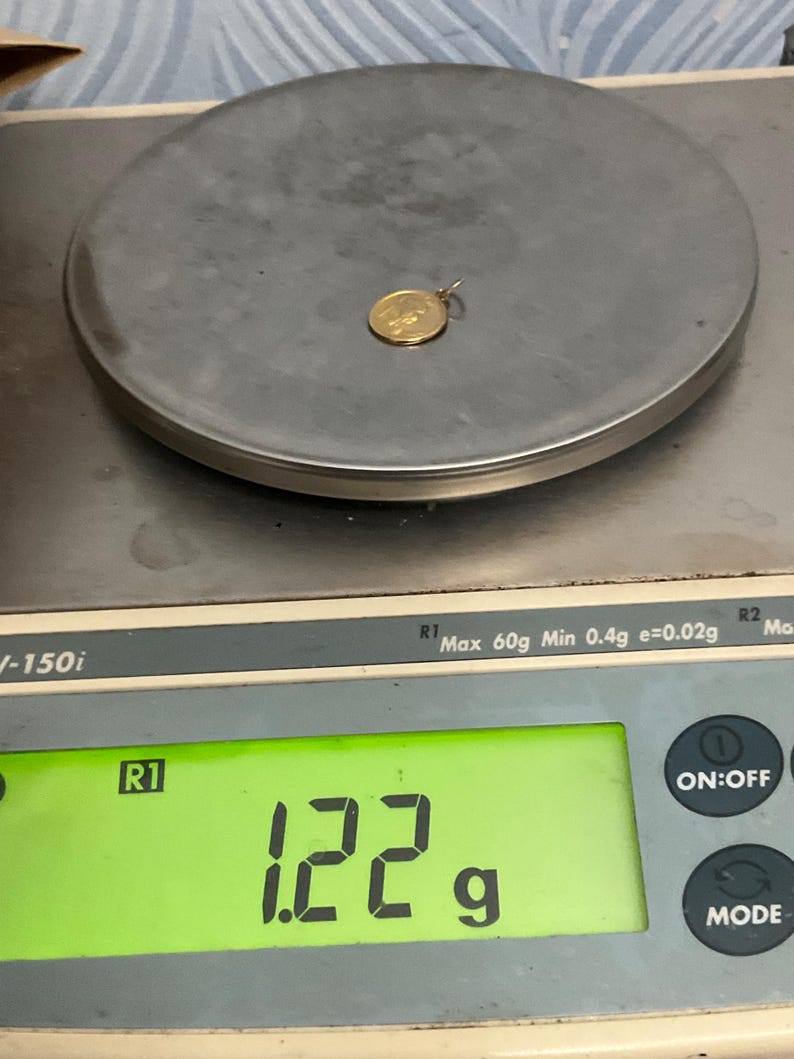 K&ouml;nnte beinhalten: Ein kleiner, runder, goldfarbener Anh&auml;nger mit gepr&auml;gtem Design, der auf einer digitalen Waage liegt. Das Display der Waage zeigt ein Gewicht von 1,22 Gramm an, wobei eine maximale Kapazit&auml;t von 60 Gramm und ein Minimum von 0,4 Gramm sichtbar sind.
