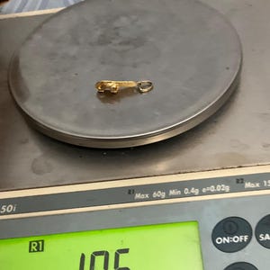 K&ouml;nnte beinhalten: Ein kleiner goldfarbener Metallanh&auml;nger mit strukturierter Oberfl&auml;che und einem runden Verbindungsring an einem Ende. Der Anh&auml;nger liegt auf einer digitalen Waage, die ein Gewicht von 1,06 Gramm anzeigt, wobei das Display den Wert deutlich wiedergibt.