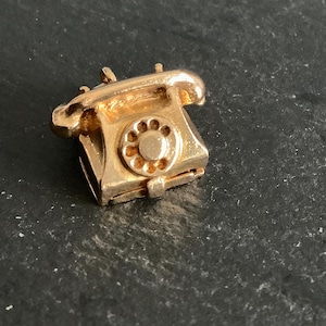 Könnte beinhalten: Ein goldfarbener Anhänger in Form eines alten Wählscheibentelefons. Der Anhänger hat ein detailliertes Design und eine kleine Schlaufe zum Anbringen an einem Armband oder einer Halskette.