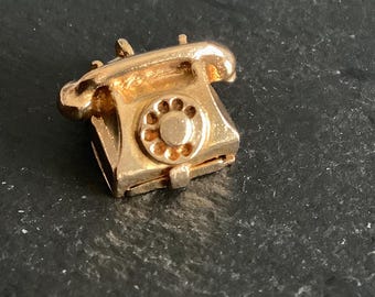 May include: A gold-colored charm in the shape of a vintage rotary phone. The charm has a detailed design and a small loop for attaching to a bracelet or necklace.