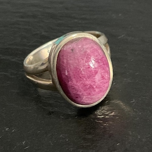 May include: A silver ring featuring a large, oval-shaped, polished magenta-pink gemstone set in a smooth metal bezel. The band of the ring is split into two distinct silver segments that connect at the base, providing a layered appearance to the design.