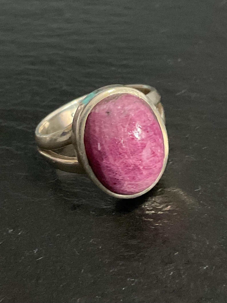 Pu&ograve; includere: Un anello in argento con una grande pietra preziosa ovale di colore rosa magenta lucido, incastonata in una montatura in metallo liscio. La fascia dell'anello &egrave; divisa in due segmenti d'argento distinti che si uniscono alla base.