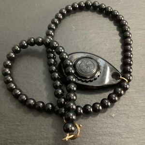 May include: A black beaded necklace with a decorative pendant. The necklace features small, round beads and a gold-colored clasp. The pendant is a dark, teardrop shape with a floral design.