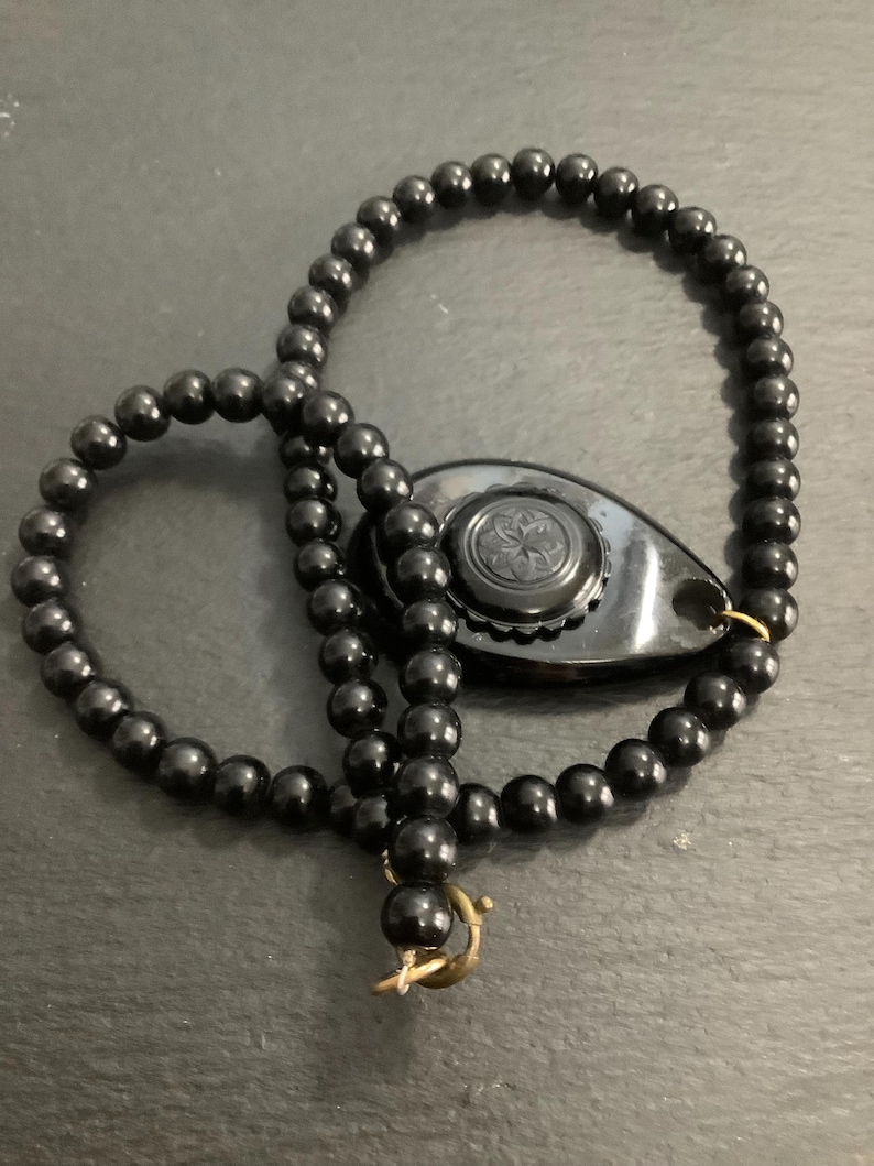 May include: A black beaded necklace with a decorative pendant. The necklace features small, round beads and a gold-colored clasp. The pendant is a dark, teardrop shape with a floral design.