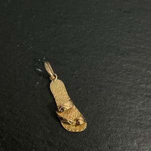K&ouml;nnte beinhalten: Ein goldener Anh&auml;nger in Form einer einzelnen Sandale mit texturiertem Fu&szlig;bett und zwei glatten Riemen auf der Oberseite. Der Anh&auml;nger verf&uuml;gt &uuml;ber einen kleinen runden Verbindungsring an der Oberseite, polierte Oberfl&auml;chen und Details.