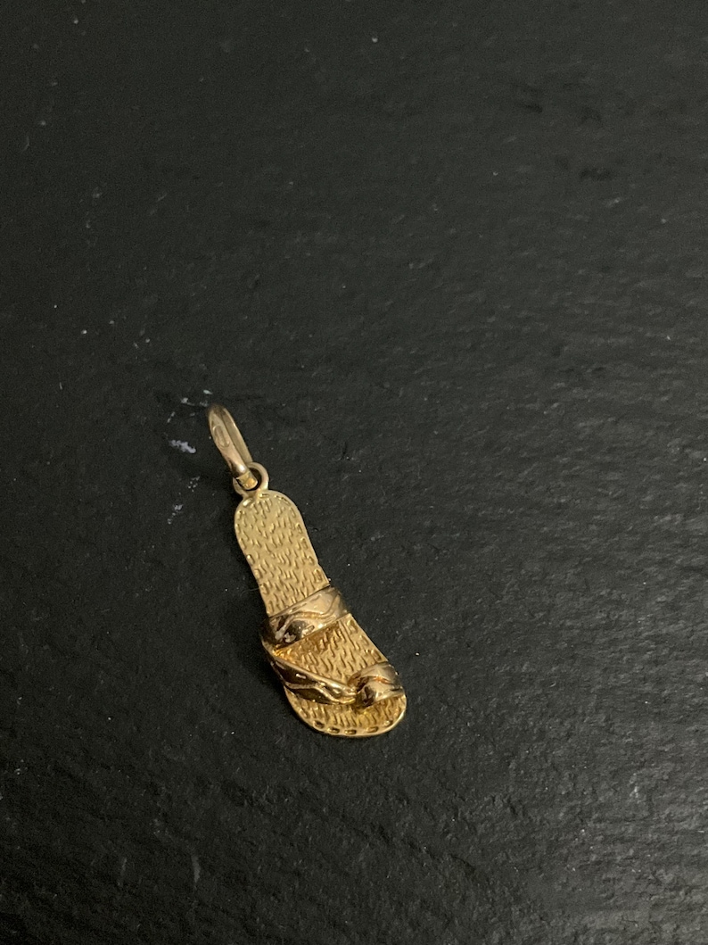 K&ouml;nnte beinhalten: Ein goldener Anh&auml;nger in Form einer einzelnen Sandale mit texturiertem Fu&szlig;bett und zwei glatten Riemen auf der Oberseite. Der Anh&auml;nger verf&uuml;gt &uuml;ber einen kleinen runden Verbindungsring an der Oberseite, polierte Oberfl&auml;chen und Details.