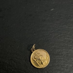 K&ouml;nnte beinhalten: Ein runder, goldfarbener Anh&auml;nger mit einem erhabenen Reliefdesign, das eine Figur zeigt, die ein Kind auf der Schulter tr&auml;gt, erg&auml;nzt durch einen kleinen Verbindungsring an der Oberseite zur Befestigung an einer Kette oder einem Band.