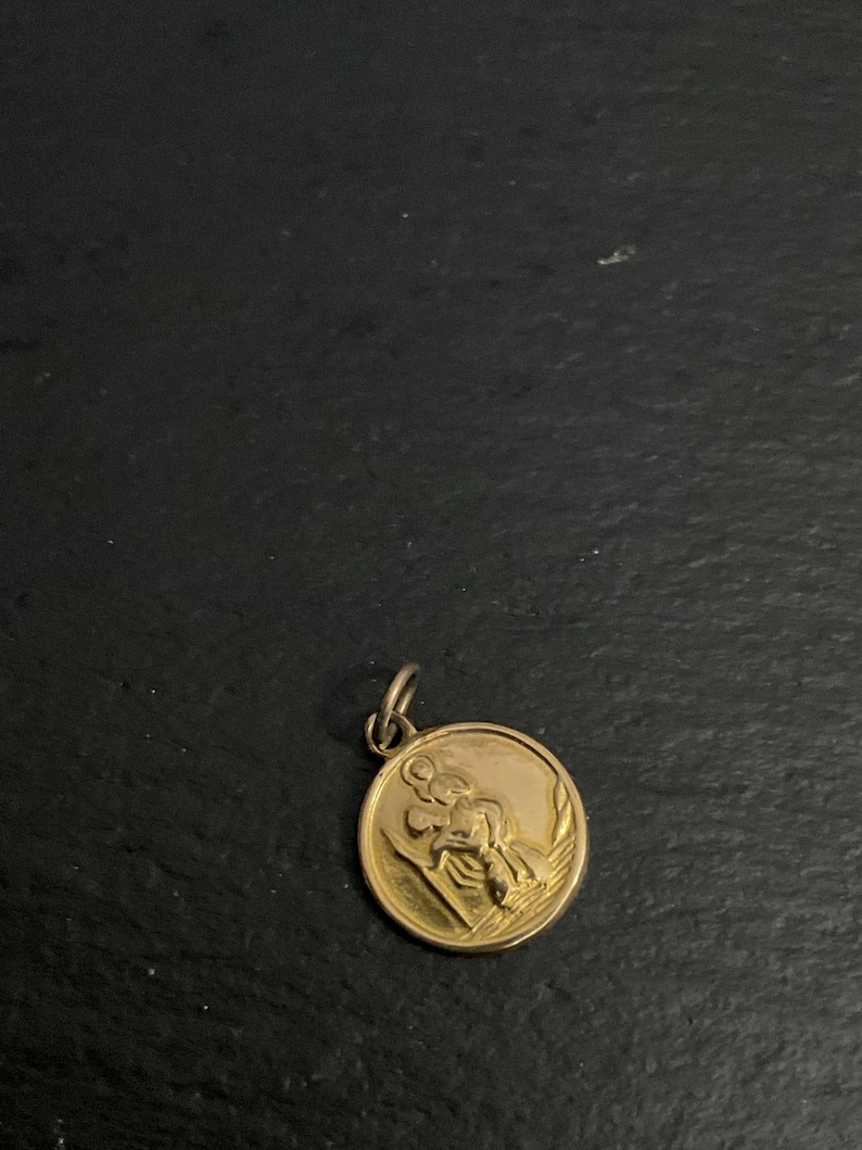 K&ouml;nnte beinhalten: Ein runder, goldfarbener Anh&auml;nger mit einem erhabenen Reliefdesign, das eine Figur zeigt, die ein Kind auf der Schulter tr&auml;gt, erg&auml;nzt durch einen kleinen Verbindungsring an der Oberseite zur Befestigung an einer Kette oder einem Band.