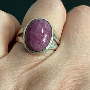 Pu&ograve; includere: Un anello in argento con una grande gemma cabochon di forma ovale color magenta intenso. La pietra ha una superficie liscia e lucida con sottili texture fibrose interne. La fascia in metallo si divide in due sezioni sottili presso la montatura.