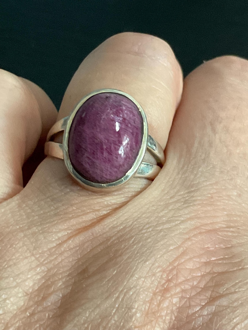 Pu&ograve; includere: Un anello in argento con una grande gemma cabochon di forma ovale color magenta intenso. La pietra ha una superficie liscia e lucida con sottili texture fibrose interne. La fascia in metallo si divide in due sezioni sottili presso la montatura.