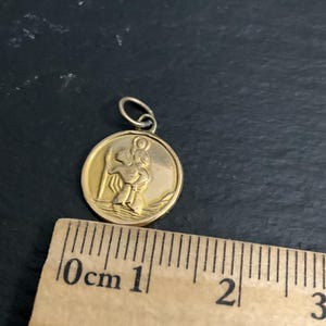 K&ouml;nnte beinhalten: Ein runder goldfarbener Anh&auml;nger mit einem Relief einer Figur, die ein Kind auf der Schulter tr&auml;gt, platziert &uuml;ber einem Holzlineal, das zeigt, dass der Anh&auml;nger einen Durchmesser von etwa 1,9 cm hat, inklusive der &Ouml;se.