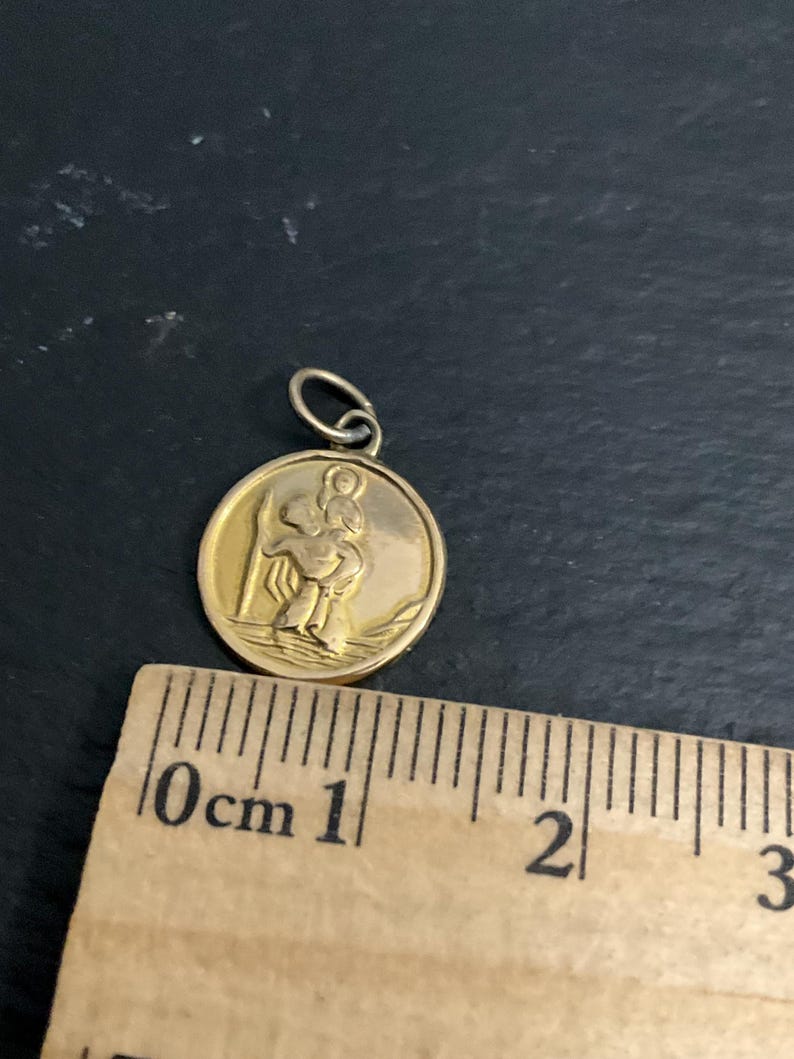 K&ouml;nnte beinhalten: Ein runder goldfarbener Anh&auml;nger mit einem Relief einer Figur, die ein Kind auf der Schulter tr&auml;gt, platziert &uuml;ber einem Holzlineal, das zeigt, dass der Anh&auml;nger einen Durchmesser von etwa 1,9 cm hat, inklusive der &Ouml;se.