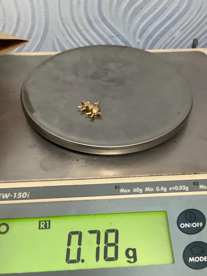 K&ouml;nnte beinhalten: Ein kleiner goldfarbener Schweineanh&auml;nger auf einer digitalen Waage. Das Display der Waage zeigt ein Gewicht von 0,78 Gramm an. Die Waage ist ein silberfarbenes Modell mit einer runden Wiegefl&auml;che und einem gr&uuml;nen Digitaldisplay.