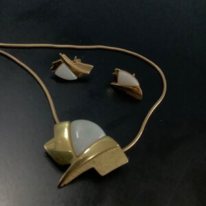 Op de afbeelding: Een gouden ketting met een witte en gouden hanger en bijpassende oorbellen. De hanger is een geometrische vorm met een witte steen in het midden.