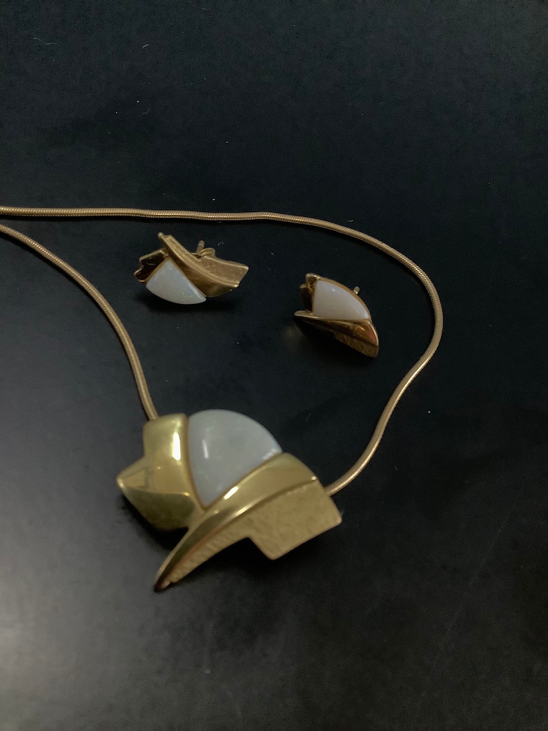 Op de afbeelding: Een gouden ketting met een witte en gouden hanger en bijpassende oorbellen. De hanger is een geometrische vorm met een witte steen in het midden.