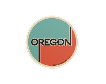 May include: A round sticker with a teal and orange background. The word "OREGON" is printed in black letters across the sticker. The text "DandWeElements.com" is printed in small black letters at the bottom of the sticker.