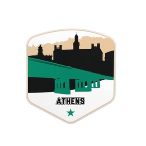 May include: A white shield-shaped sticker with a black silhouette of a city skyline and a green bridge. The text "ATHENS" is printed in black at the bottom of the sticker with a green star.