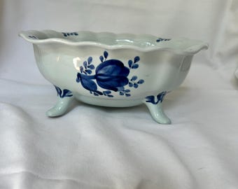 May include: A white ceramic bowl with a scalloped rim and three small feet. The bowl is decorated with blue floral designs. The bowl is approximately 8 inches in diameter.