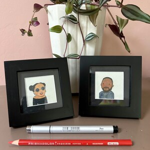 May include: Two black picture frames with white mats, each containing a hand-drawn portrait of a person. The frames are on a light brown surface with a red pencil and a silver marker.