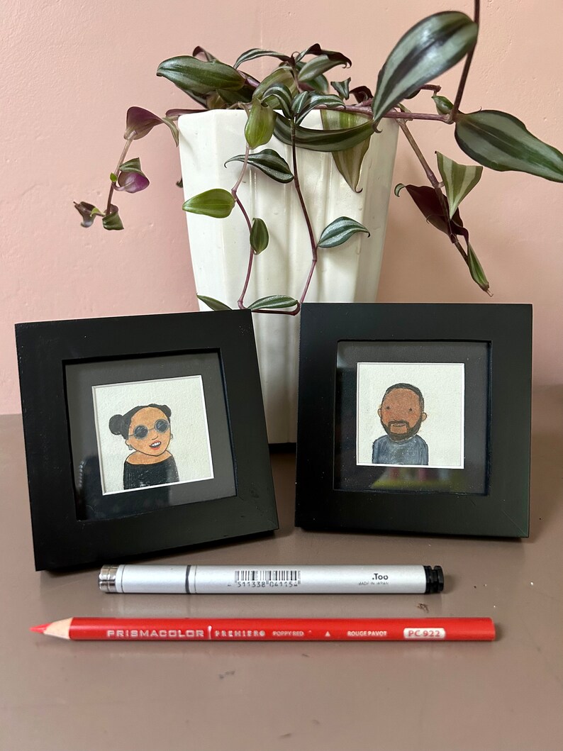 May include: Two black picture frames with white mats, each containing a hand-drawn portrait of a person. The frames are on a light brown surface with a red pencil and a silver marker.