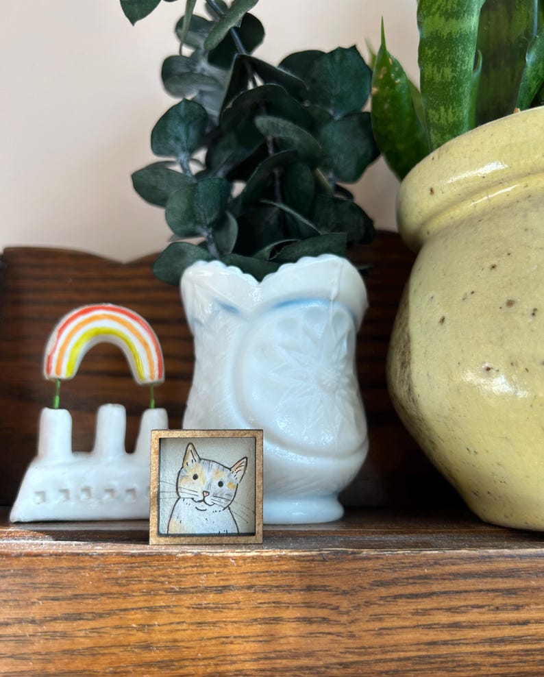 May include: A small, square wooden frame with a watercolor painting of a cat's face. The cat has orange and gray markings. A rainbow decoration and a white vase are also visible.