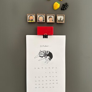 May include: A refrigerator with decorative magnets and a calendar. The magnets include fruit shapes and framed portraits. The calendar, held by a red clip, displays the month of October with a cat illustration and the text "peopleyoumaymeet.com".