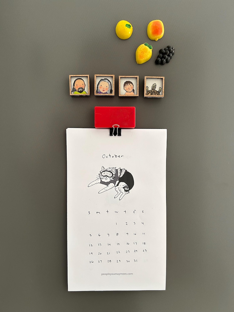 May include: A refrigerator with decorative magnets and a calendar. The magnets include fruit shapes and framed portraits. The calendar, held by a red clip, displays the month of October with a cat illustration and the text "peopleyoumaymeet.com".