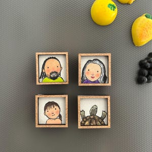 May include: Four small, square wooden magnets with hand-drawn portraits and a turtle. The portraits depict a man, a woman, and a child. The fourth magnet features a turtle. Yellow and orange fruit magnets are also visible.