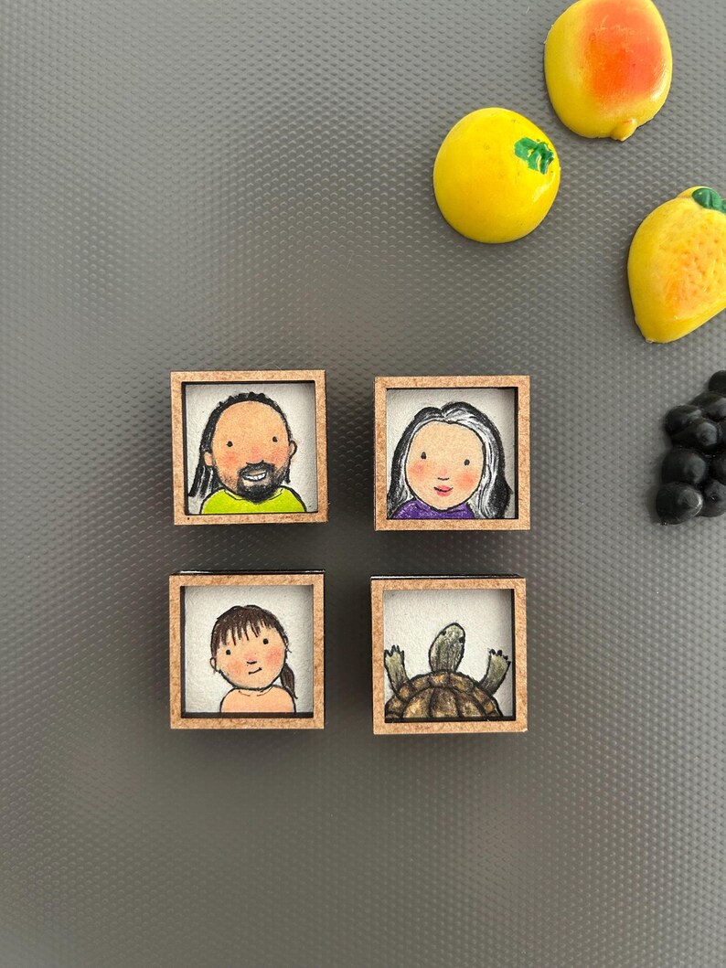 May include: Four small, square wooden magnets with hand-drawn portraits and a turtle. The portraits depict a man, a woman, and a child. The fourth magnet features a turtle. Yellow and orange fruit magnets are also visible.