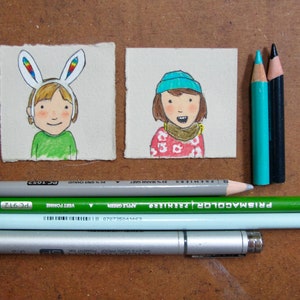 May include: Two hand-drawn illustrations on paper cards. The first card shows a person with blonde hair wearing a rainbow bunny ear headband. The second card shows a person with brown hair wearing a blue beanie and a red and white patterned sweater.  The cards are surrounded by colored pencils and a silver pen.