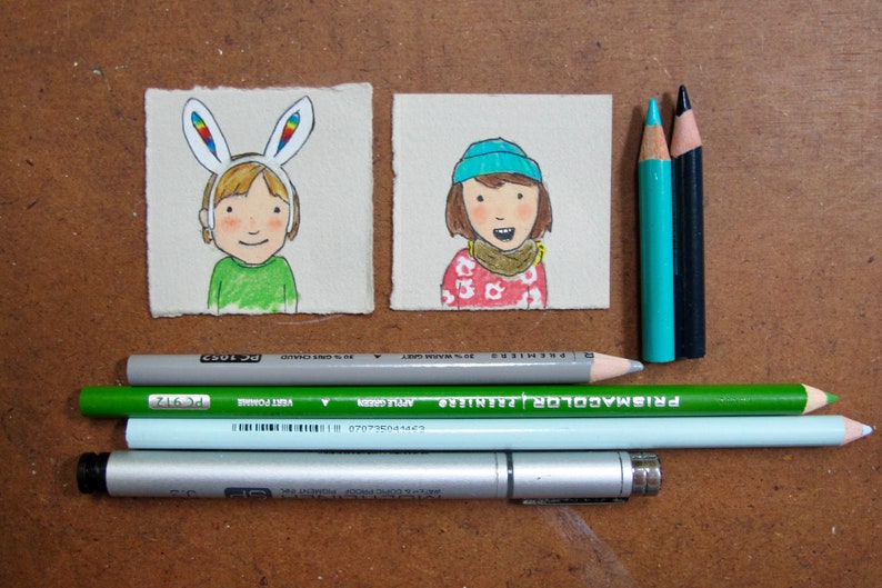 May include: Two hand-drawn illustrations on paper cards. The first card shows a person with blonde hair wearing a rainbow bunny ear headband. The second card shows a person with brown hair wearing a blue beanie and a red and white patterned sweater.  The cards are surrounded by colored pencils and a silver pen.