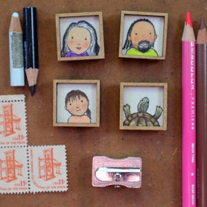 May include: A collection of miniature framed illustrations, including portraits and a turtle, alongside colored pencils, stamps, and a pencil sharpener. The stamps read "USA 11" and "FREEDOM OF THE PRESS". The pencils are pink, brown, and blue.