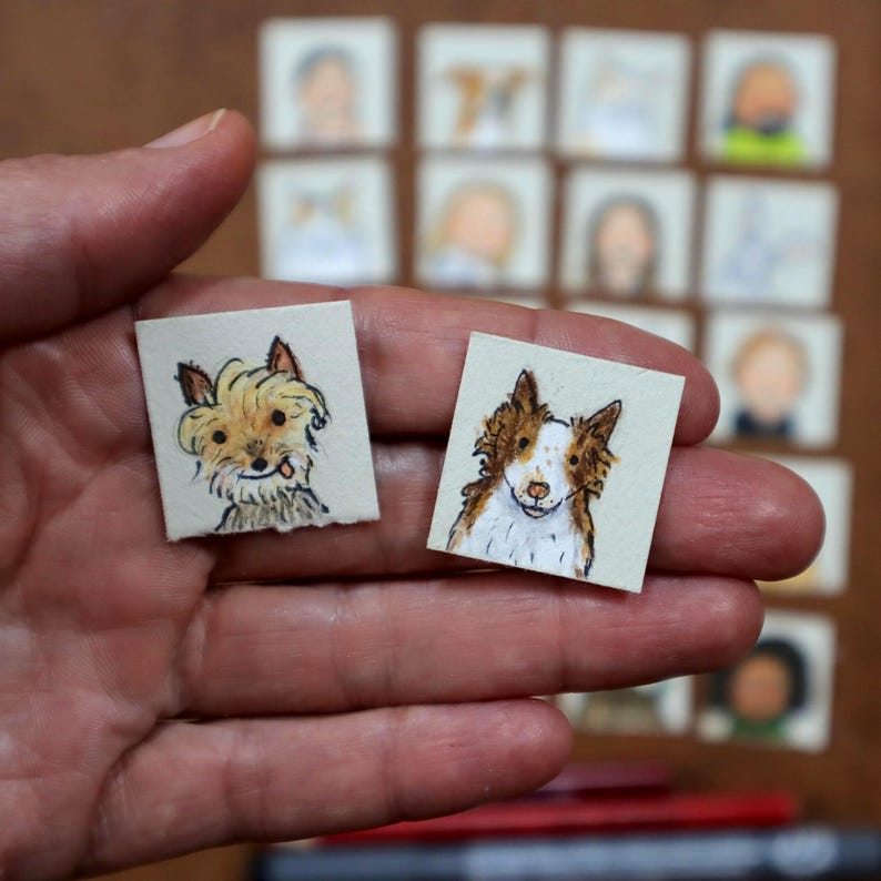 May include: Two small, square watercolor paintings of dogs are held in a hand. One dog is a tan and white terrier mix, and the other is a brown and white border collie. The background features other small, square paintings of people.
