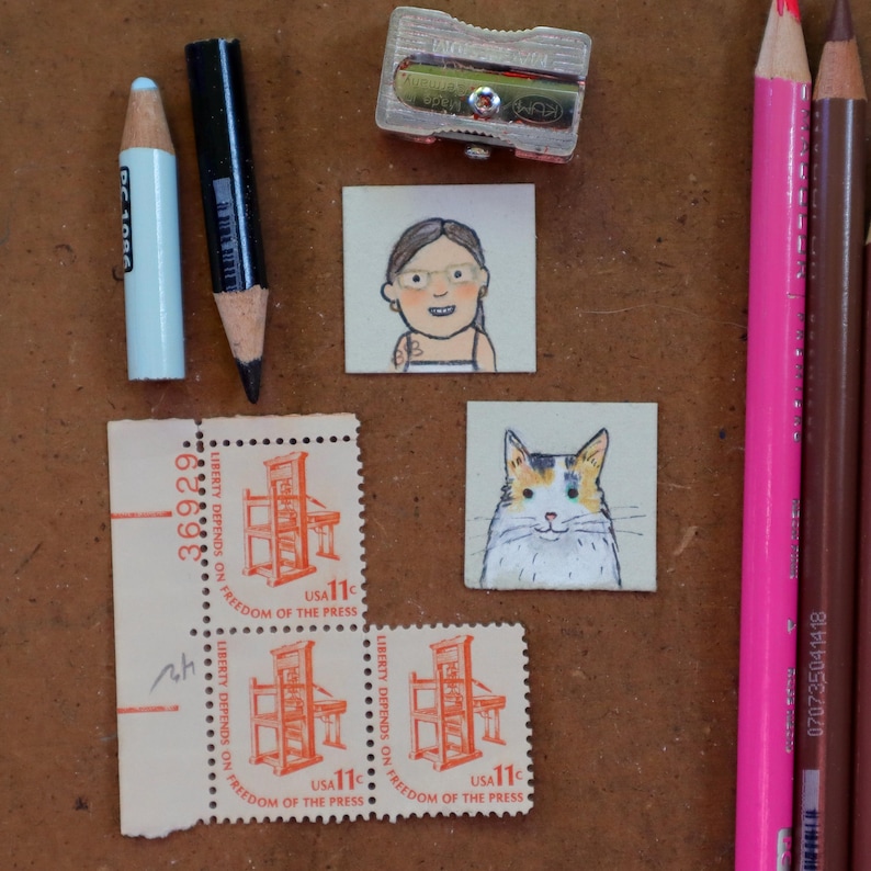 May include: A collection of art supplies and small artworks on a brown surface. Includes a light blue pencil, a black pencil, a pencil sharpener, two small drawings, and two "Freedom of the Press" stamps.