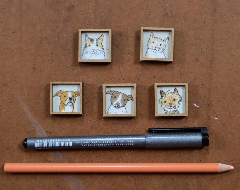 May include: Five small, square wooden framed pet portraits: two cats and three dogs. A silver pen and orange pencil are below the portraits. The cat portraits are of a calico cat and a white and orange cat. The dog portraits are of a brown and white dog, a brown and white dog, and a tan dog.