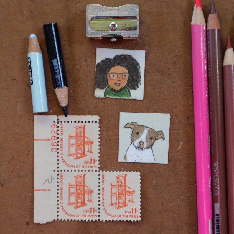 May include: An assortment of art supplies and miniature artwork on a brown surface. Includes colored pencils, a pencil sharpener, miniature portraits, and vintage postage stamps with the text "LIBERTY DEPENDS ON FREEDOM OF THE PRESS".