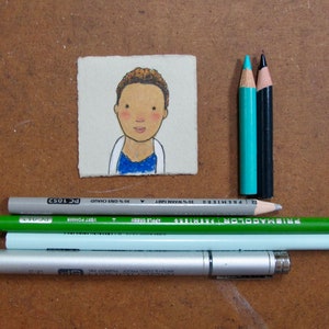May include: A small, hand-painted illustration of a person with brown hair and a blue shirt on a white background. The illustration is mounted on a piece of cardboard. There are also several colored pencils and a silver pen on a brown surface.
