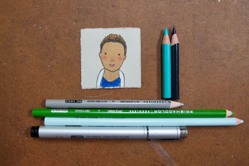May include: A small, hand-painted illustration of a person with brown hair and a blue shirt on a white background. The illustration is mounted on a piece of cardboard. There are also several colored pencils and a silver pen on a brown surface.