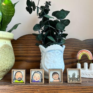 May include: Four small framed art pieces with portraits and a turtle illustration. The frames are wood. A white vase with green leaves and a ceramic rainbow decor are also visible. The items are displayed on a wooden shelf.