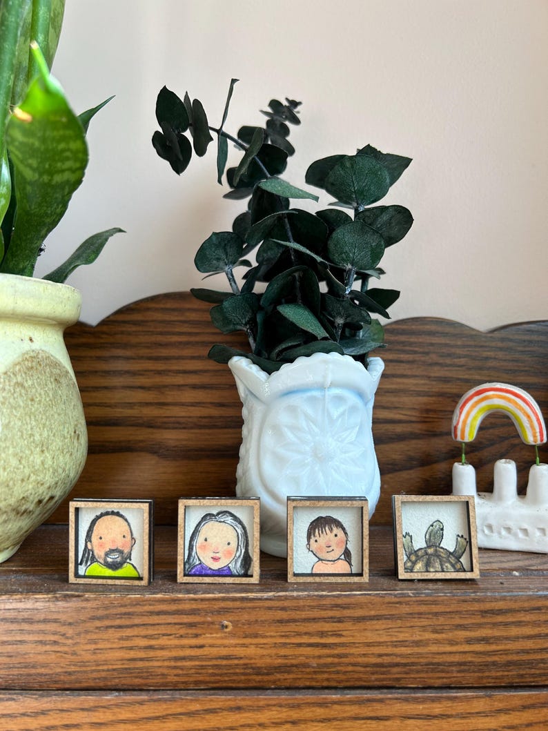 May include: Four small framed art pieces with portraits and a turtle illustration. The frames are wood. A white vase with green leaves and a ceramic rainbow decor are also visible. The items are displayed on a wooden shelf.