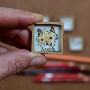 May include: A miniature artwork featuring a hand-drawn portrait of a dog within a small, square wooden frame. The dog has light brown fur, dark eyes, and a small tongue sticking out. The artwork is held in a person's hand.
